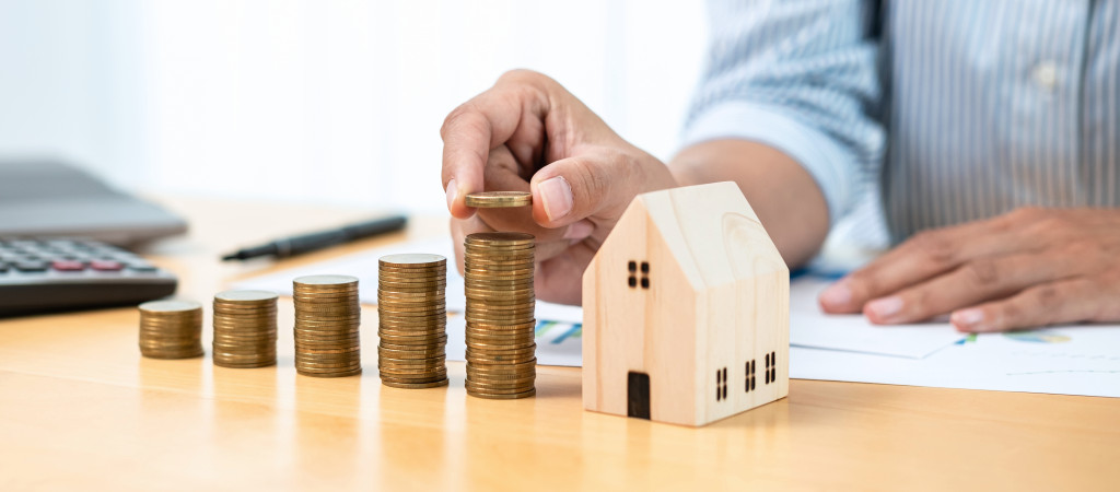 A person stacking coins in a rising order on a table with a miniature wooden house